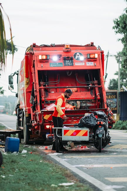 A street scene featuring a red rubbish collection vehicle in the background, with its rear compaction area open and a worker standing beside it, actively loading waste materials. The worker is wearing
